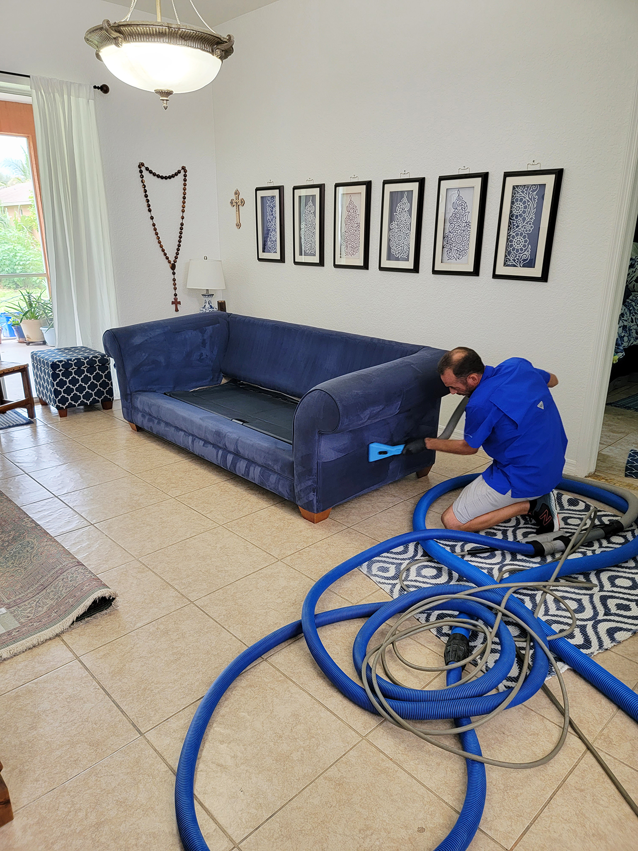 Professional upholstery cleaning in progress, technician using hot water extraction on blue couch, surrounded by home decor and cleaning hoses.